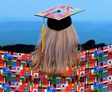 Photo of woman wearing graduation mortarboard and an international sash representing multiple countries looking out across a mountain landscape.